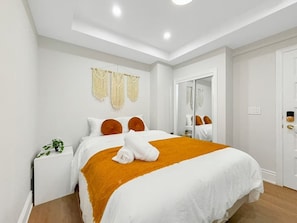 Uma cama bem arrumada com um edredom branco e laranja, uma pequena mesa de cabeceira com uma planta e um espelho na parede.