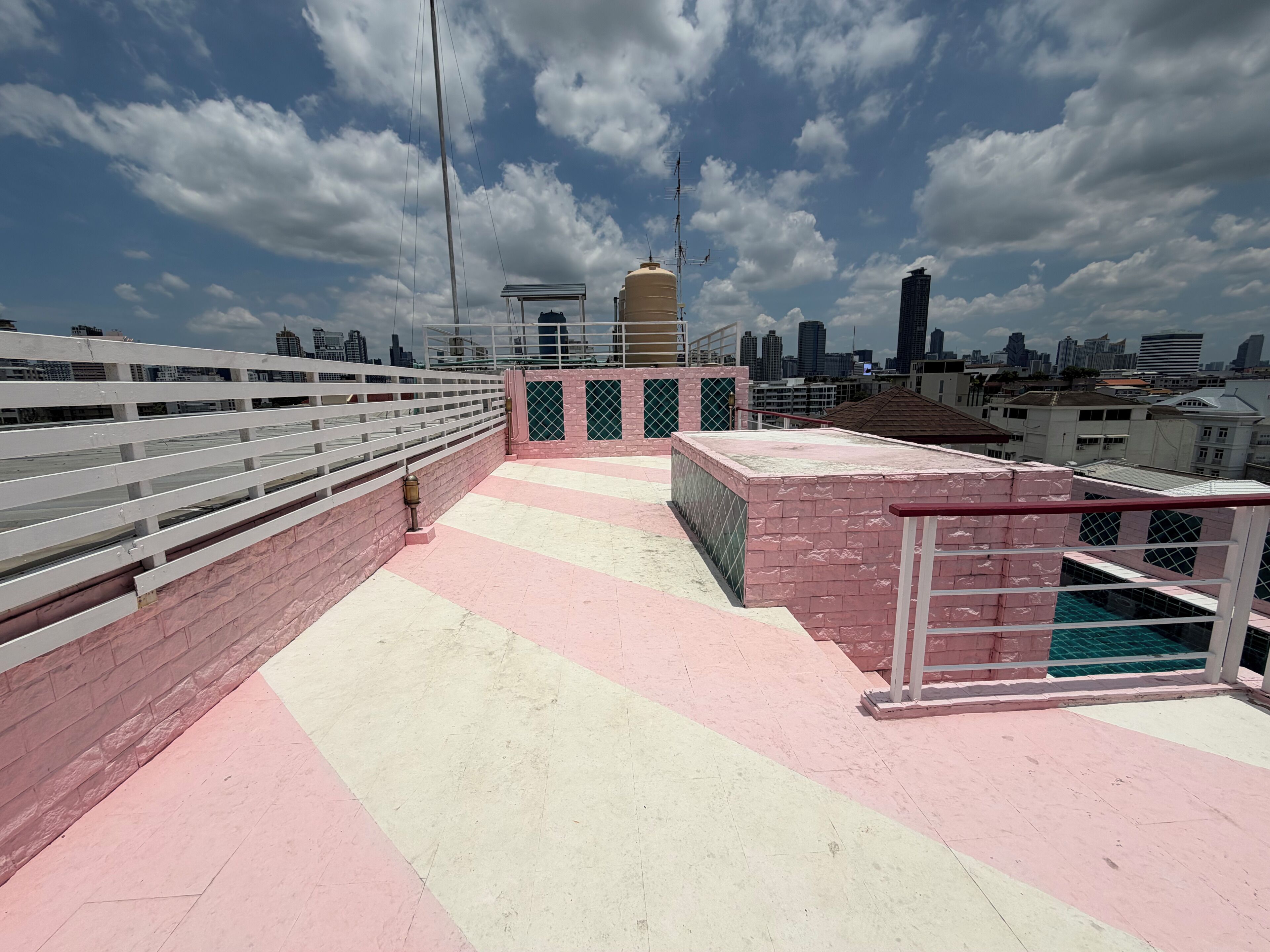 Una terraza en la azotea con barandilla blanca, suelo rosa y beige, y un horizonte urbano de fondo.