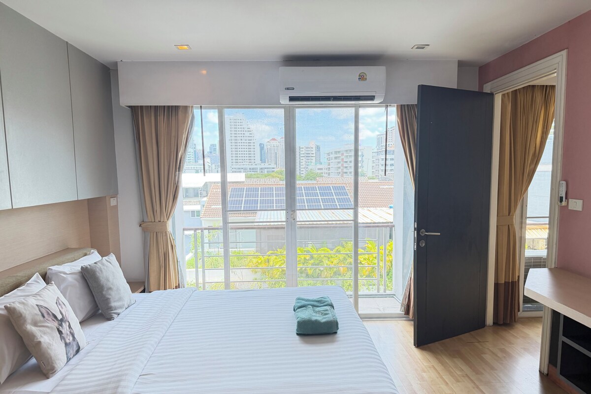 A hotel room with a bed, curtains, an air conditioning unit, a view of buildings and greenery, and a door.