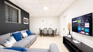 A modern living room with a grey sofa, a dining table with chairs, and a flat-screen TV mounted on the wall displaying various streaming services.