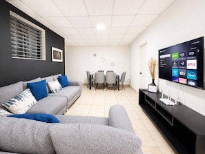A modern living room with a grey sofa, a dining table with chairs, and a flat-screen TV mounted on the wall displaying various streaming services.