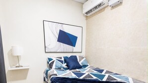 A bedroom with a bed, a wall-mounted air conditioner, a framed abstract painting, and a bedside lamp.
