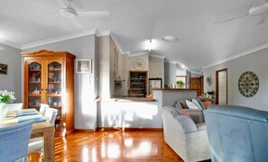 A spacious living room with a wooden floor, a sofa, a dining table with chairs, and a kitchen area in the background. - Summit House - Elevated Family Retreat (Mackay)