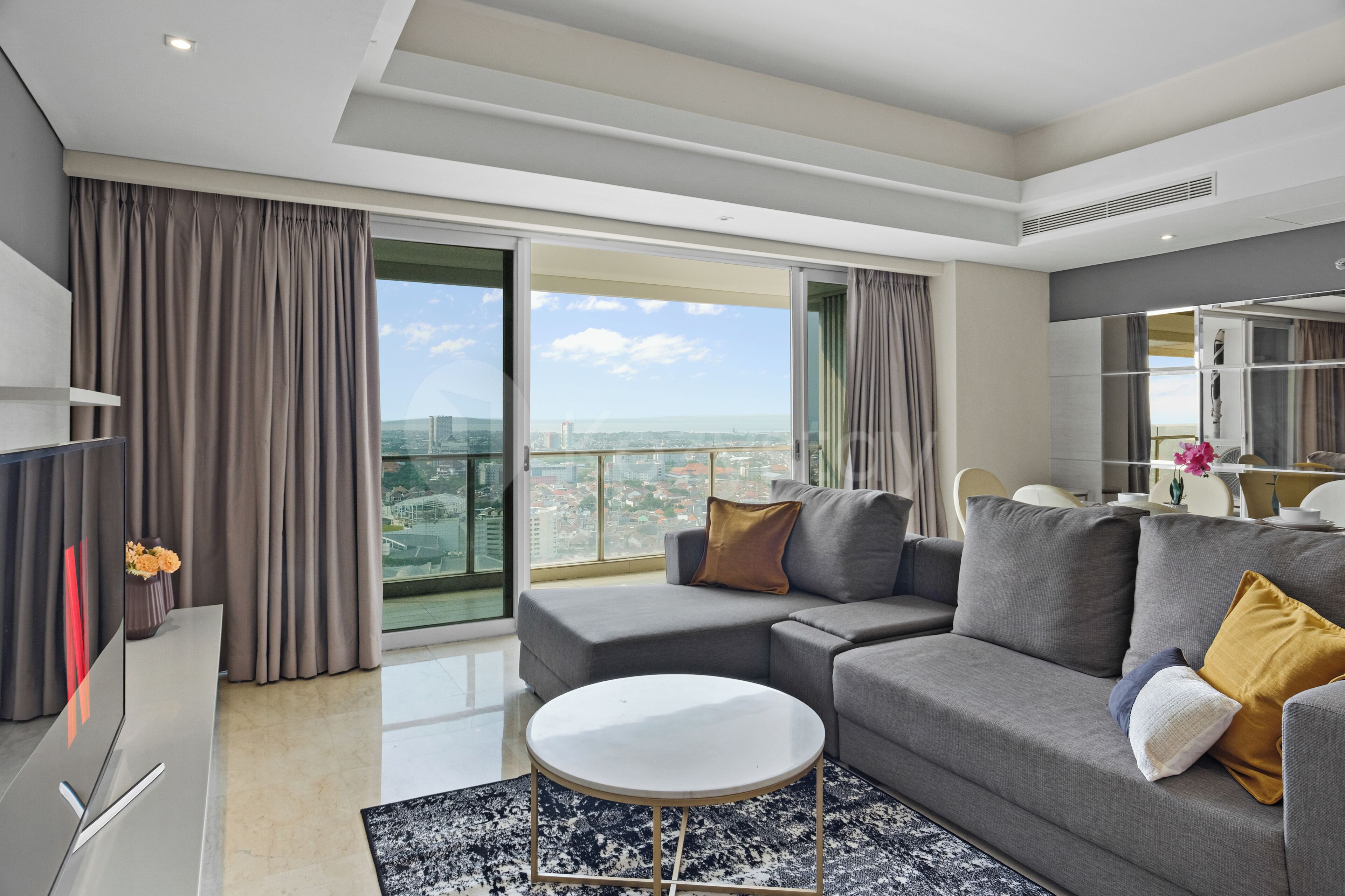 A modern living room with a grey sofa, a round coffee table, and a view of the cityscape through large windows.