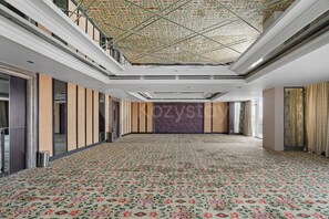 Ein geräumiger Saal mit gemustertem Teppich, einer hohen Decke mit geometrischem Muster und einem großen Fenster auf der rechten Seite.