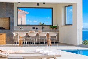 A modern outdoor kitchen with a large window overlooking a clear sky and a pool. - Villa rOvni, Labin, Croatia (Labin)