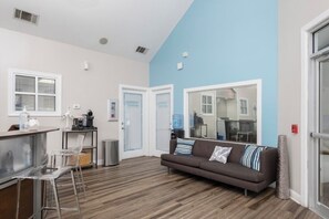 A modern living room with a brown sofa, clear bar stools, a coffee machine, and a kitchen area with a window and a door. - Gainesville 3 Ensuite Bedrooms Near UF Shands Free Pool Gym Parking and WiFi (Gainesville)