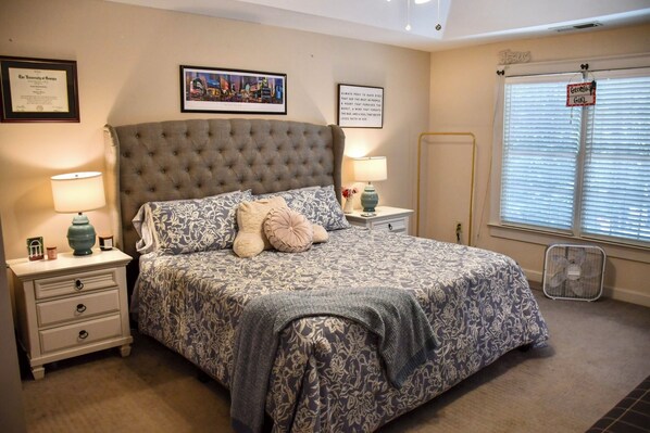 A bedroom with a tufted headboard, a large bed with floral bedding, two bedside tables with lamps, a framed certificate on the wall, and a window with blinds.