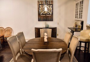 A dining room with a round table, six chairs, a decorative wall piece above the table, and a window with blinds.