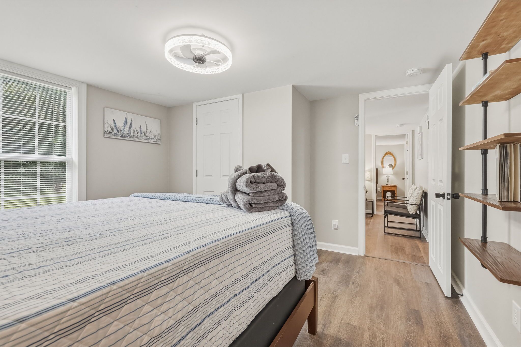 A bedroom with a bed, a bedside lamp, a window with blinds, and a door leading to another room.