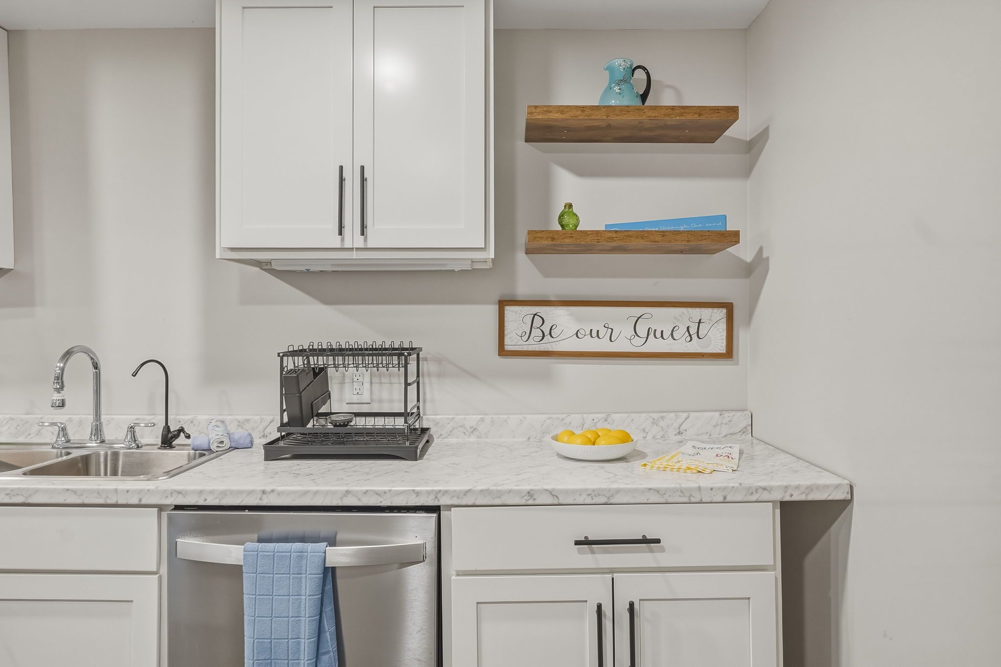 A kitchen with white cabinets, a stainless steel sink, a dish rack, a kettle, and a bowl of lemons on the counter.