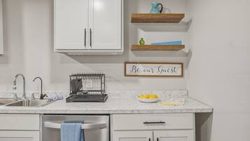 A kitchen with white cabinets, a stainless steel sink, a dish rack, a kettle, and a bowl of lemons on the counter.