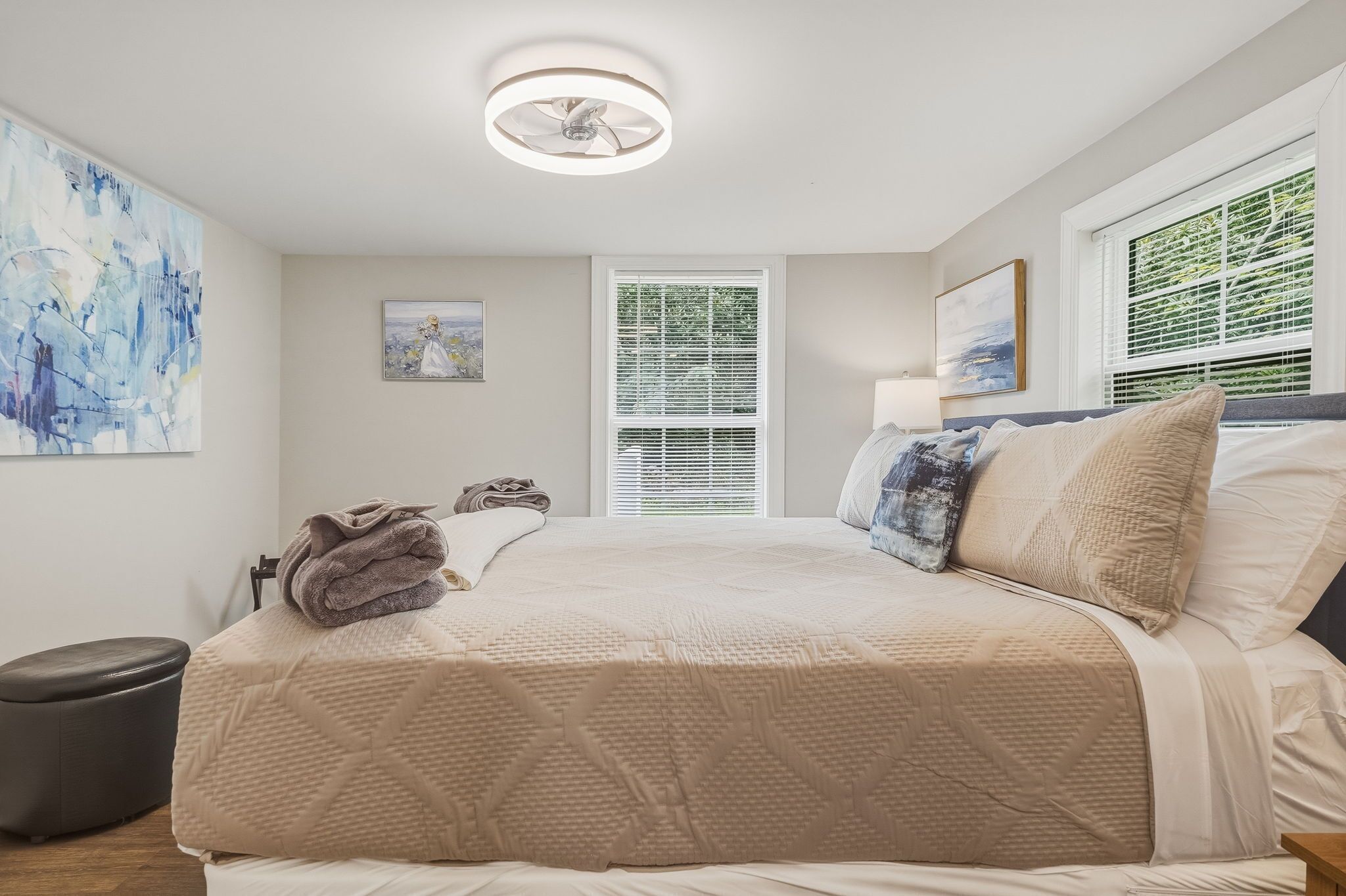 A bedroom with a bed, a bedside table, a lamp, a window with blinds, and artwork on the walls.