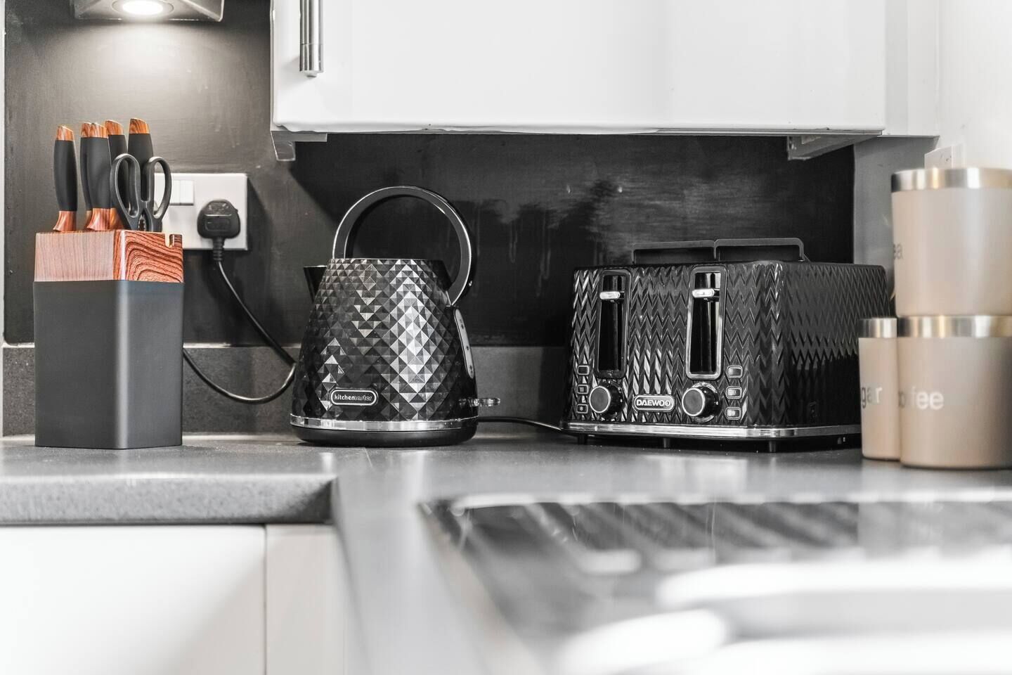 A kitchen counter with a coffee maker, a kettle, and a toaster. There are also utensils in a holder and a coffee machine on the wall.