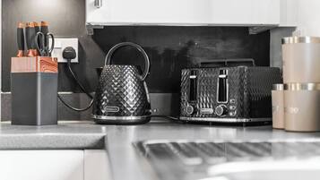 A kitchen counter with a coffee maker, a kettle, and a toaster. There are also utensils in a holder and a coffee machine on the wall.