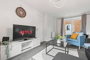 Un salón moderno con un televisor de pantalla plana mostrando 'NETFLIX', una puerta blanca, un sofá azul con un cojín amarillo, y una pared de ladrillo visible a través de una puerta de cristal.