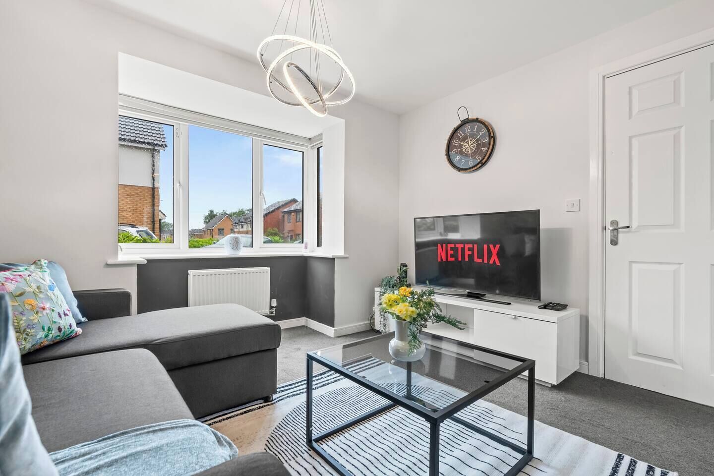 A modern living room with a sofa, a glass coffee table, a television displaying 'NETFLIX', a wall clock, and a window with a view of houses.