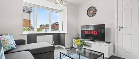 Ein modernes Wohnzimmer mit einer Couch, einem Glastisch, einem Fernseher, der 'NETFLIX' zeigt, einer Wanduhr und einem Fenster mit Blick auf Häuser.
