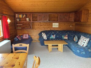 A cozy cabin interior with a wooden ceiling, a blue sofa with patterned cushions, a wooden table, and a bookshelf with books. - Chalet Glimuschir Appartement/Fewo, Dusche, WC Appartement/Fewo, Dusche, WC by Interhome (Rosswald)