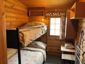 Quarto de cabana de madeira compacto com beliches, uma mesinha e janela com cortinas.