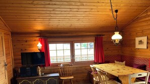 A wooden cabin interior with a dining table, chairs, a television, and a window with curtains. - Chalet Glimuschir Appartement/Fewo, Dusche, WC Appartement/Fewo, Dusche, WC by Interhome (Rosswald)