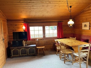 A wooden cabin interior with a dining table, chairs, a television, and a window with curtains. - Chalet Glimuschir Appartement/Fewo, Dusche, WC Appartement/Fewo, Dusche, WC by Interhome (Rosswald)