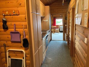 Puinen mökki, jonka sisätiloissa on keittiöalue, johon kuuluu kaappeja, jääkaappi ja ruokapöytä tuoleineen.