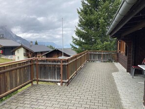 A wooden deck with a brick floor, surrounded by a wooden fence, and overlooking a mountainous landscape. - Chalet Glimuschir Appartement/Fewo, Dusche, WC Appartement/Fewo, Dusche, WC by Interhome (Rosswald)