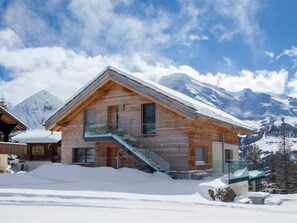 Una cabaña de madera con escalera de cristal, rodeada de nieve y montañas.