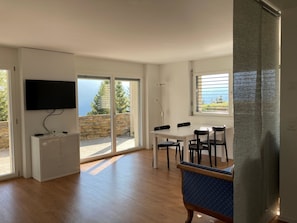 Una sala de estar moderna con un televisor de pantalla plana, un comedor con mesa y sillas, y un amplio ventanal corredizo que da a un espacio exterior.