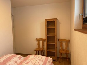 Um quarto pequeno com duas cadeiras de madeira, uma estante alta de madeira e uma janela com persianas.