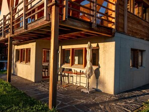 Uma cabana de madeira com um alpendre de pedra, uma sala de jantar com mesa e cadeiras, e uma área de estar coberta ao ar livre.