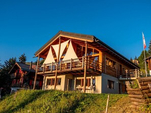 A two-story wooden chalet with a balcony, surrounded by greenery.
