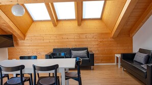 Ein Loft-Zimmer mit Holzbalkendecke, Oberlichtern, einem Essbereich mit Stühlen und Tisch sowie einer Sitzecke mit Sofa und Kissen.