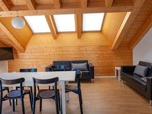 Ein Loft-Zimmer mit Holzbalkendecke, Oberlichtern, einem Essbereich mit Stühlen und Tisch sowie einer Sitzecke mit Sofa und Kissen.