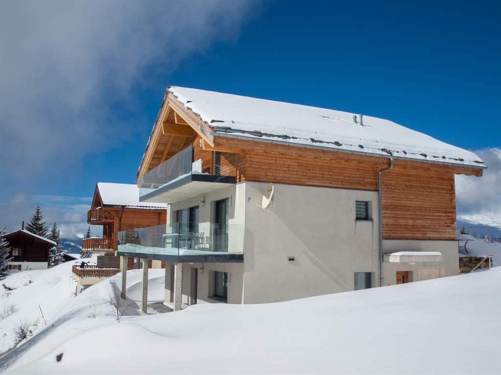 Un chalet moderno de dos pisos con fachada de madera y un balcón, rodeado de terreno nevado.