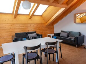 A modern loft-style room with a high ceiling, wooden beams, a skylight, a white dining table, black chairs, a grey sofa, and a small white table.