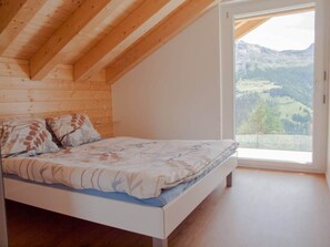 A bedroom with a wooden ceiling, a bed with floral bedding, and a large window offering a view of a mountainous landscape.
