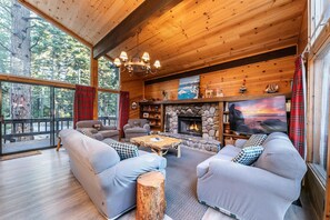 Ein gemütliches Blockhaus-Interieur mit Kamin aus Naturstein, Holzbalkendecke und großen Fenstern mit Blick auf einen Wald.