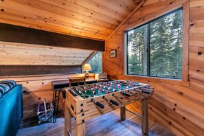 A cozy room with wooden walls, a foosball table, a blue sofa, and a window with a forest view. - Pinecone Paradise Chalet-Dog Friendly Walk to Lake (Carnelian Bay)
