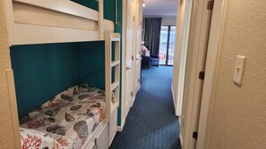 A narrow hallway with bunk beds on one side and a view of a room with a blue carpet and curtains. - The Sandbox @ Casa Del Mar Condos (Galveston)