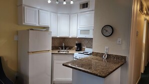 A compact kitchen with white cabinetry, a built-in microwave, a stove, and a clock on the wall. - The Sandbox @ Casa Del Mar Condos (Galveston)