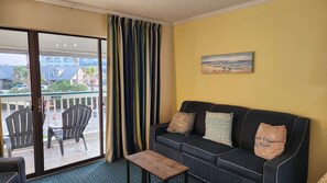 A room with a sliding glass door leading to a balcony, a black sofa with patterned cushions, and a small wooden table. - The Sandbox @ Casa Del Mar Condos (Galveston)