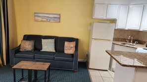 A compact living space with a sofa, a small table, a kitchen area with a refrigerator, and a painting on the wall. - The Sandbox @ Casa Del Mar Condos (Galveston)
