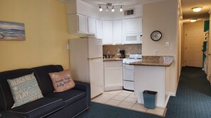 A compact kitchen with white cabinets, a microwave, and a stove. There's a black couch with decorative pillows and a small trash bin. - The Sandbox @ Casa Del Mar Condos (Galveston)
