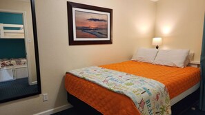 A hotel room with a bed, a framed picture on the wall, a bedside lamp, and a window with curtains. - The Sandbox @ Casa Del Mar Condos (Galveston)