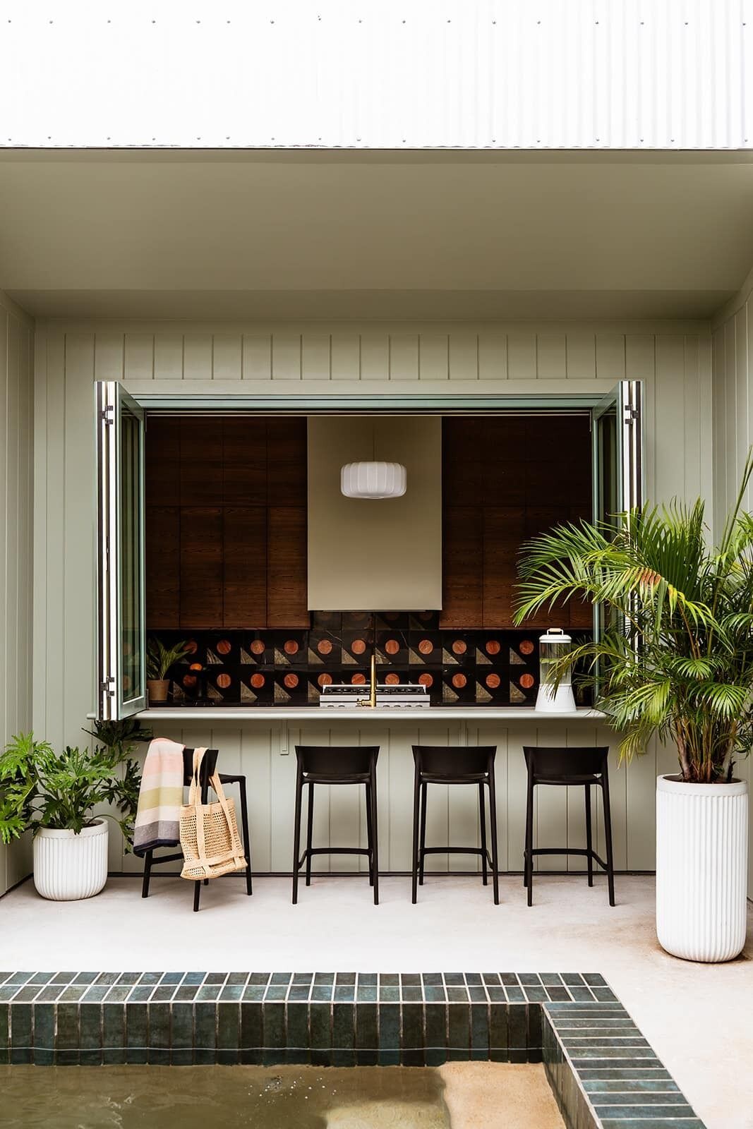 An outdoor kitchen area with a bar stool seating arrangement, a hanging light fixture, and potted plants.