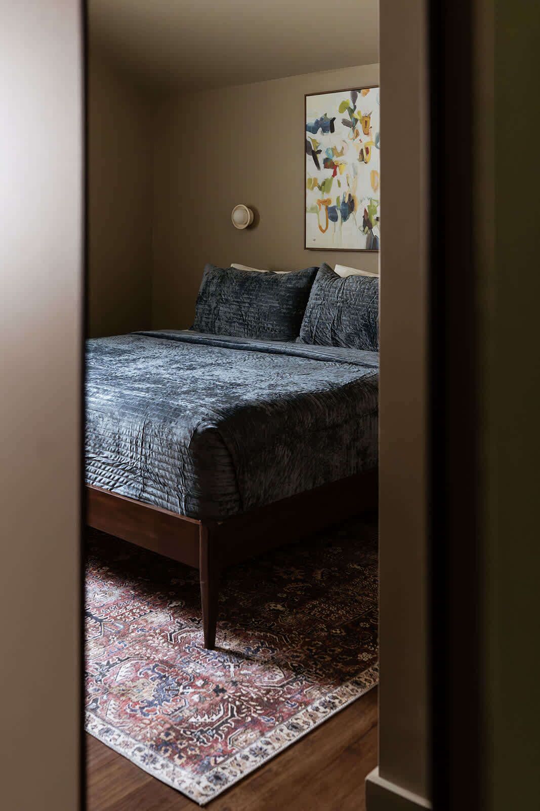 Een slaapkamer met een bed, een kamerkleed met dessin en een wandlamp.