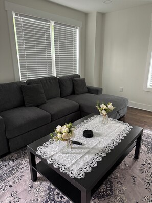 Ein Wohnzimmer mit einem dunkelgrauen Sofa, einem schwarzen Couchtisch mit weißer Spitzenauflage und einer Vase mit weißen Blumen.