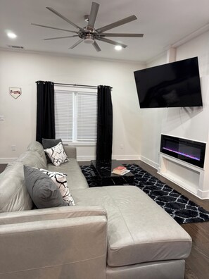 A modern living room with a sofa, a coffee table, a flat-screen TV mounted on the wall, and a ceiling fan.